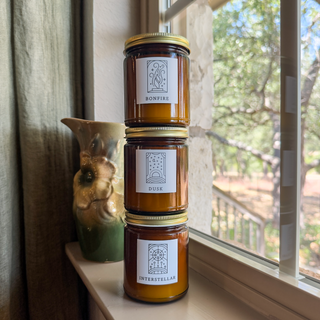 Bonfire, Dusk, and Interstellar candles stacked in amber jars beside a green ceramic pitcher and a window.