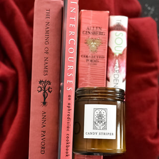 Candy Striper candle with art deco label picturing hands and herbs. Candle is shown on red blanket with four red books.